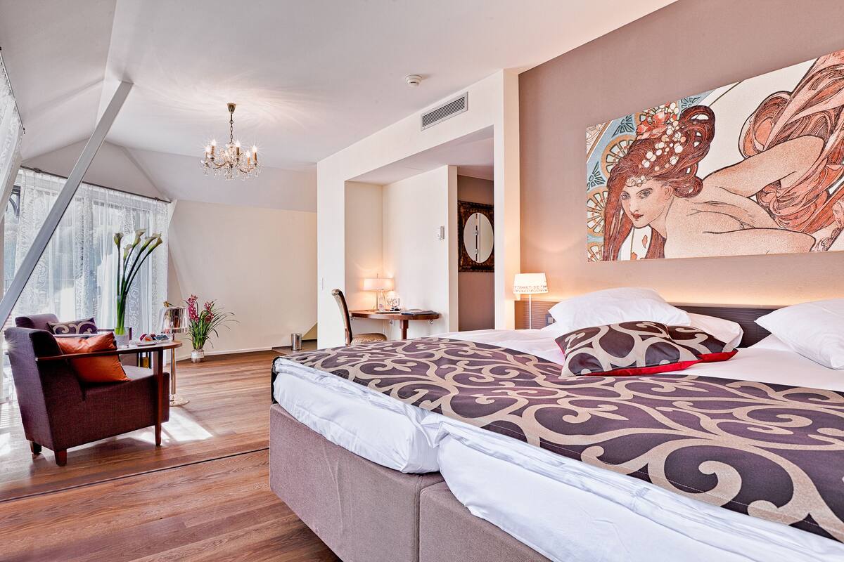 A hotel room with a large bed, a sitting area with a sofa and armchair, a desk, and a large window with curtains.