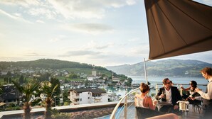 A terrace with a view of a town and lake, featuring outdoor seating and a palm tree. - Belvédère Strandhotel (Spiez)