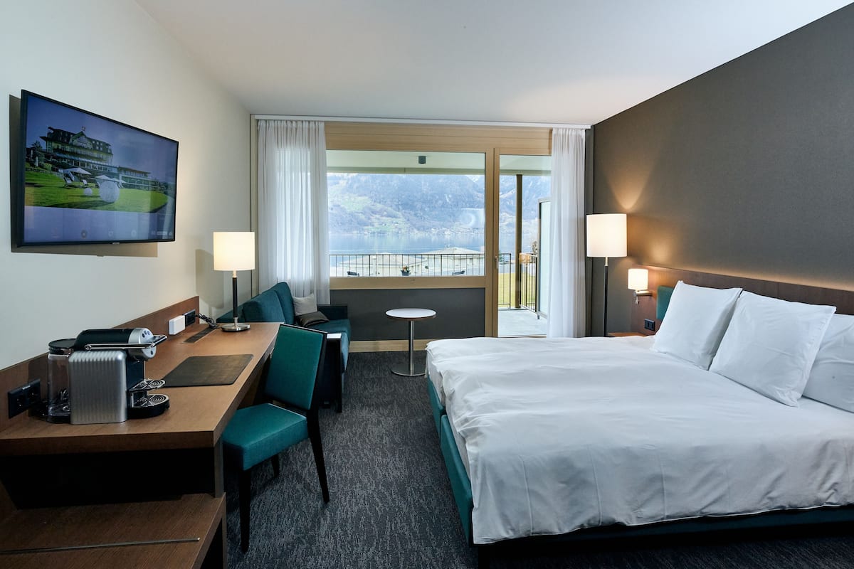 A hotel room with a bed, a desk with a computer, a chair, a small table, and a TV.