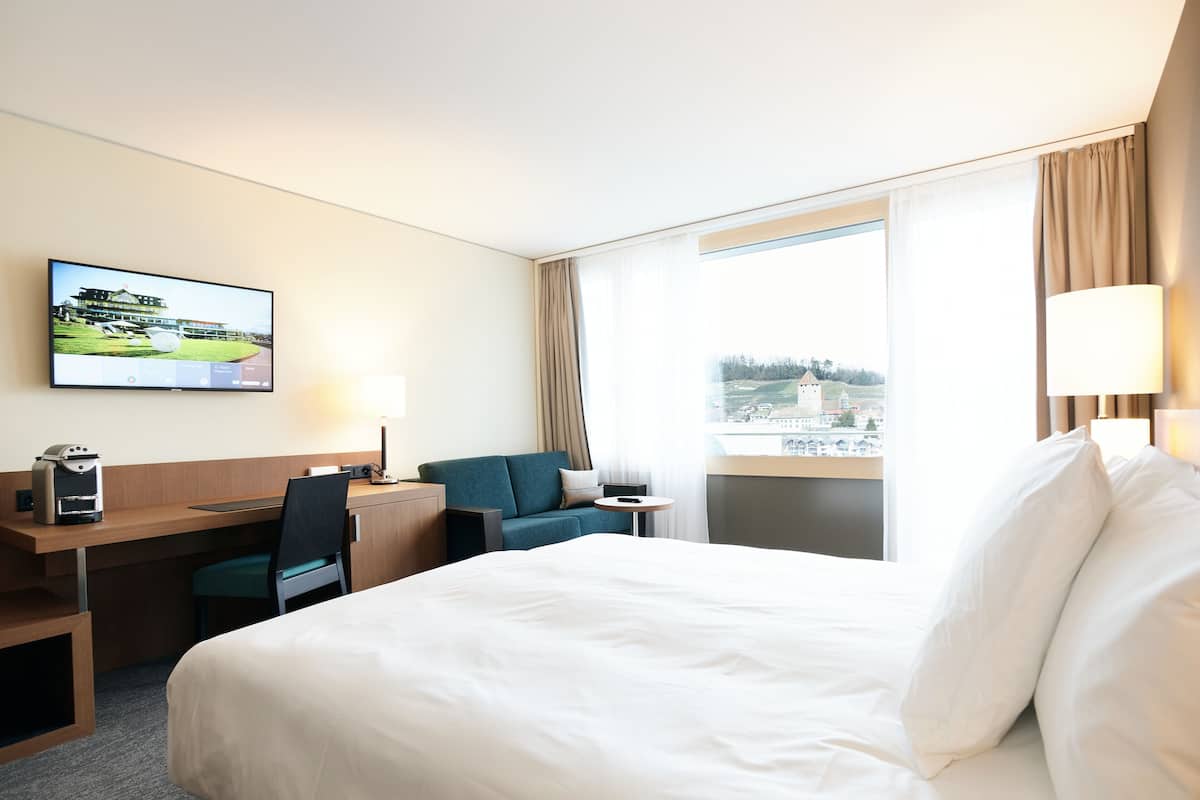 A hotel room with a large bed, a desk with a computer, a chair, a TV on the wall, and a window with a view of a building.