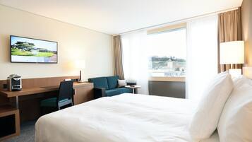 Habitación de hotel con una cama grande, un escritorio con computadora, una silla, un televisor en la pared y una ventana con vista a un edificio.