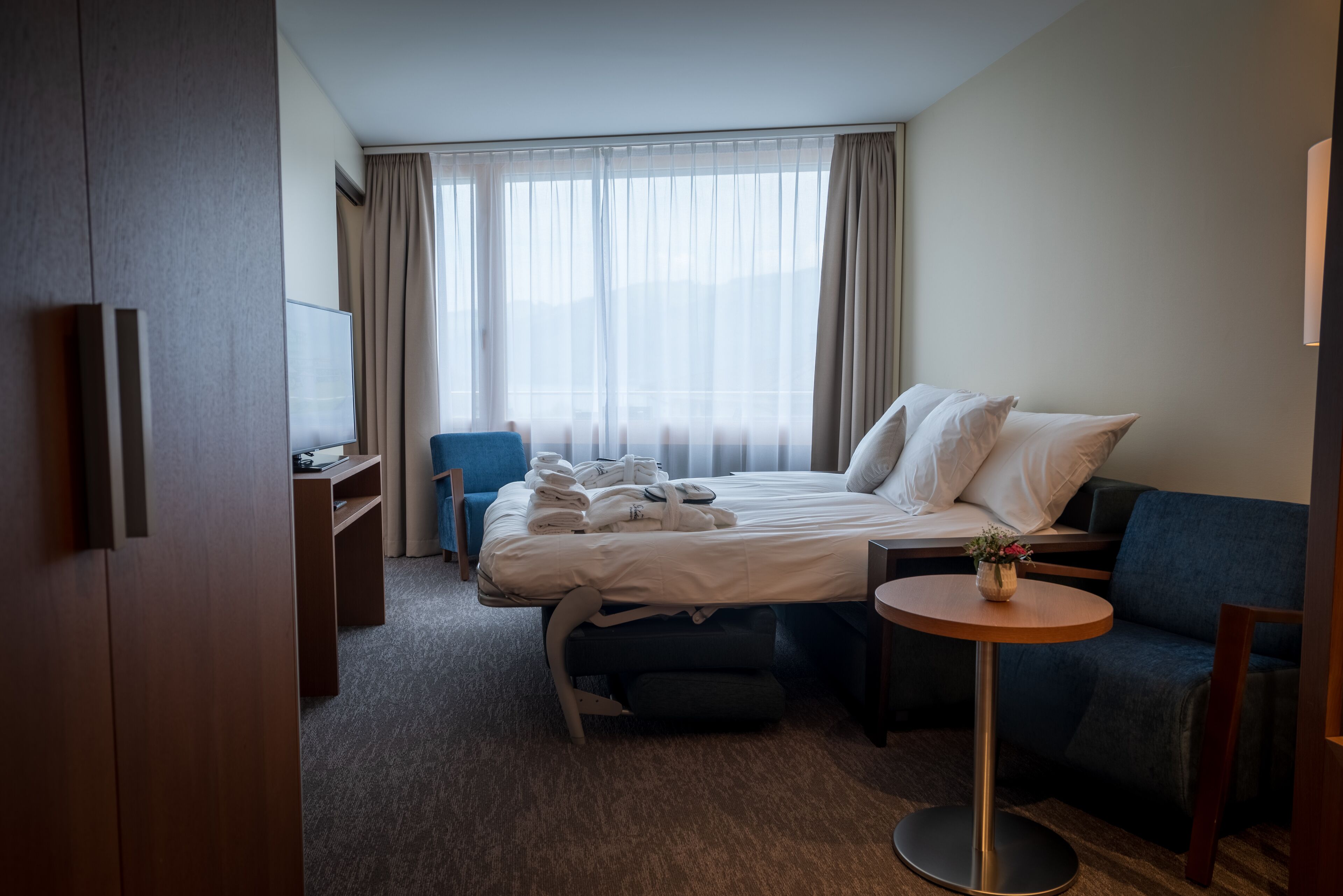 A hotel room with a bed, a desk, a chair, a sofa, and a small table with a vase.