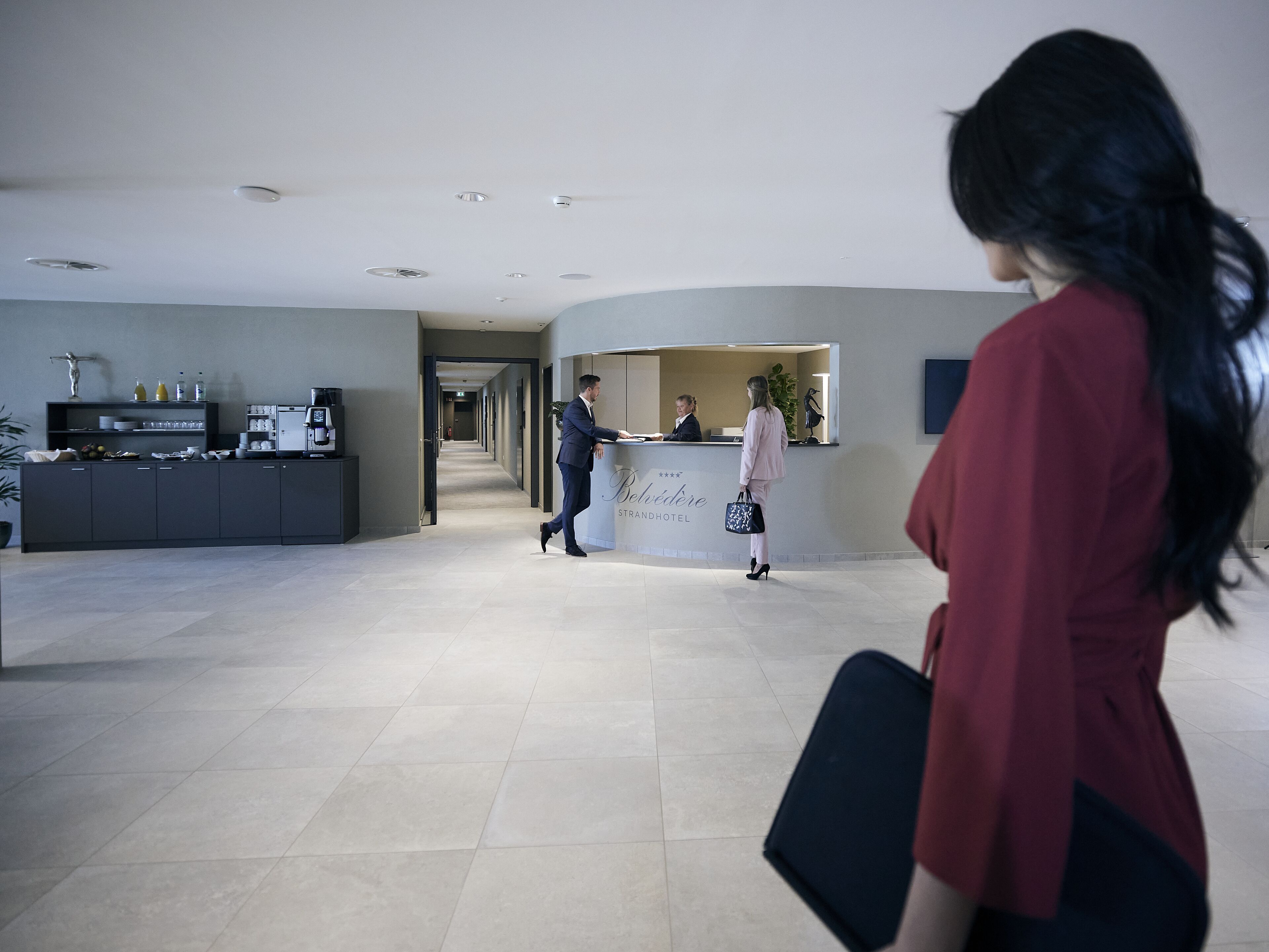 Zona de recepción de un hotel con un mostrador, un hombre con traje y una mujer con vestido rojo.