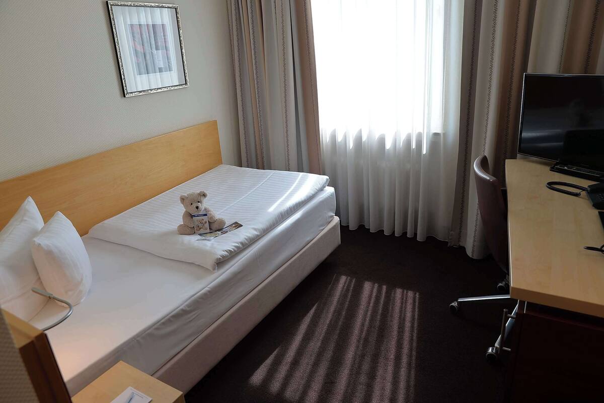 Ein Hotelzimmer mit einem Bett, einem Schreibtisch mit Computer, einem Stuhl und einem Teddybären auf dem Bett.