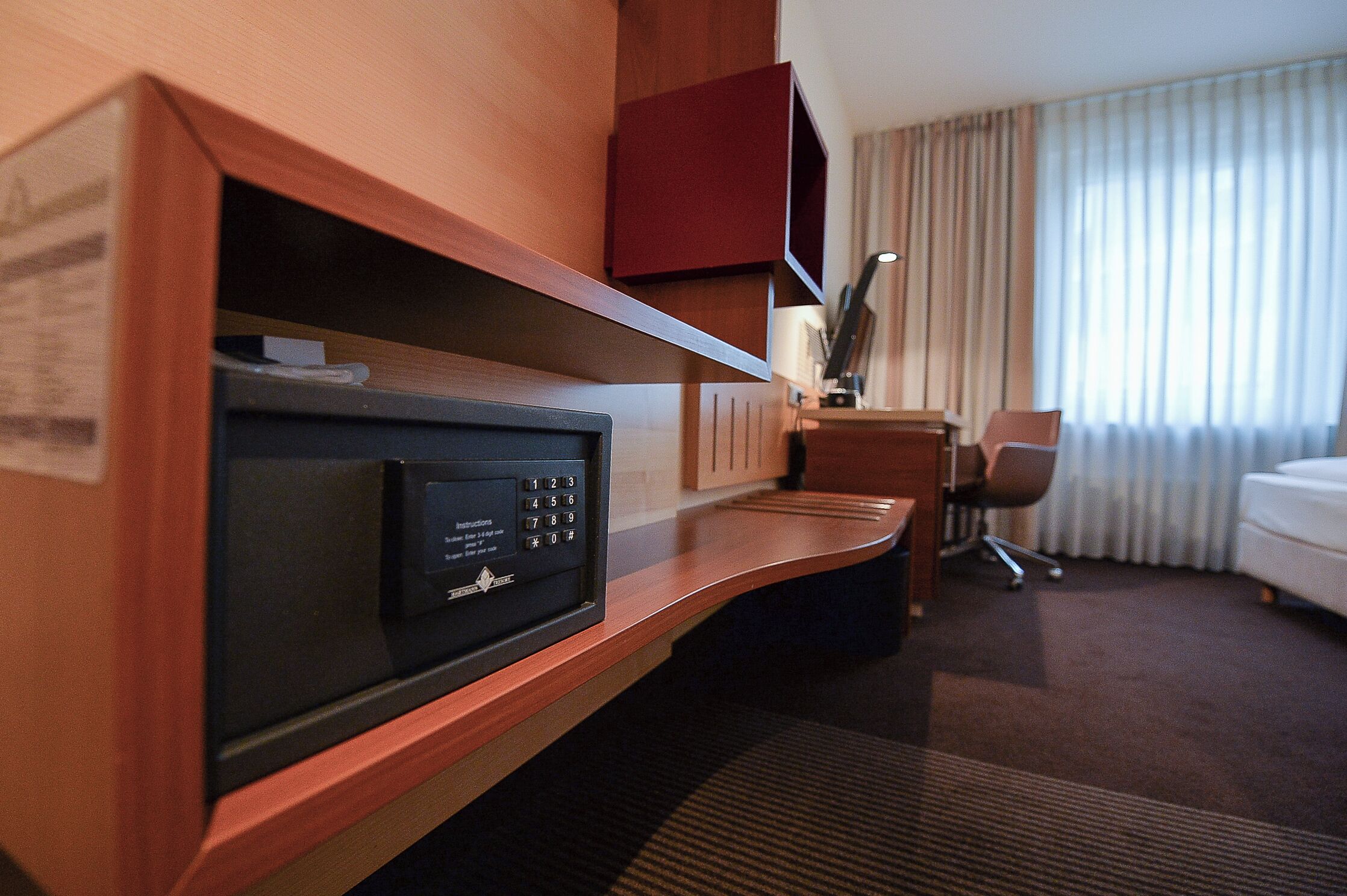 A hotel room with a built-in microwave, a desk with a lamp, a chair, and a bed.