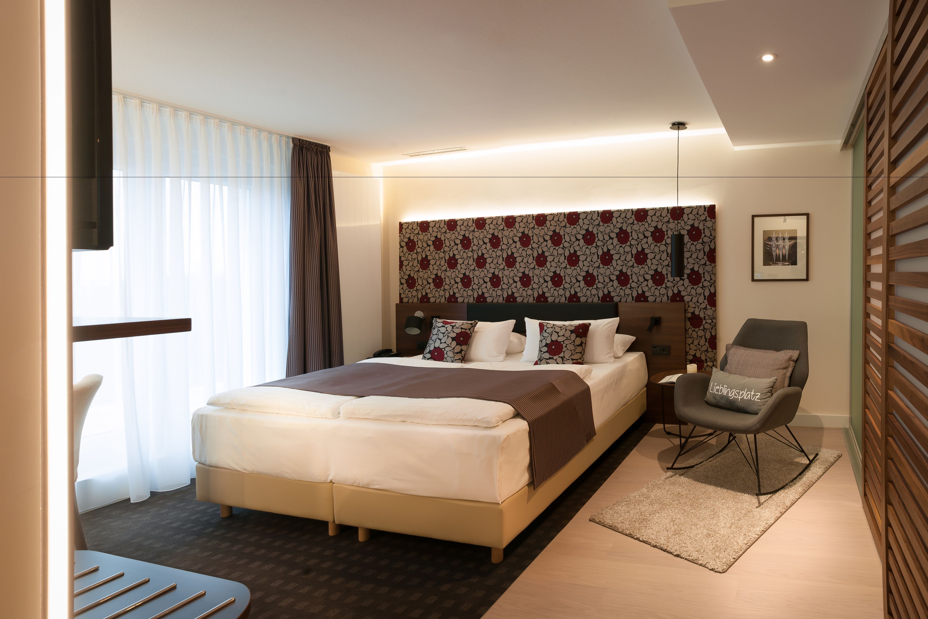 Ein modernes Hotelzimmer mit einem großen Bett, einer Sitzecke mit einem Sessel und einem gemusterten Kopfteil.