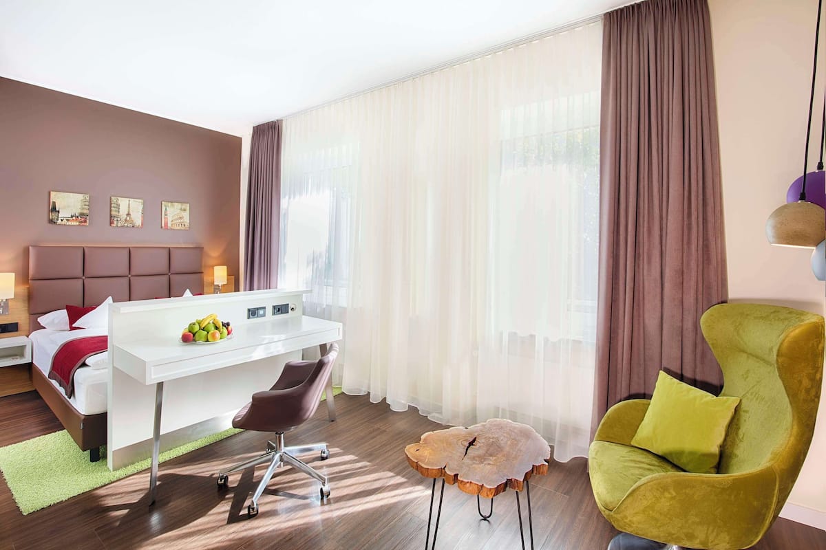 Ein modernes Hotelzimmer mit einem Bett, einem Schreibtisch, einem Stuhl, einem kleinen Hocker und einem grünen Sessel.