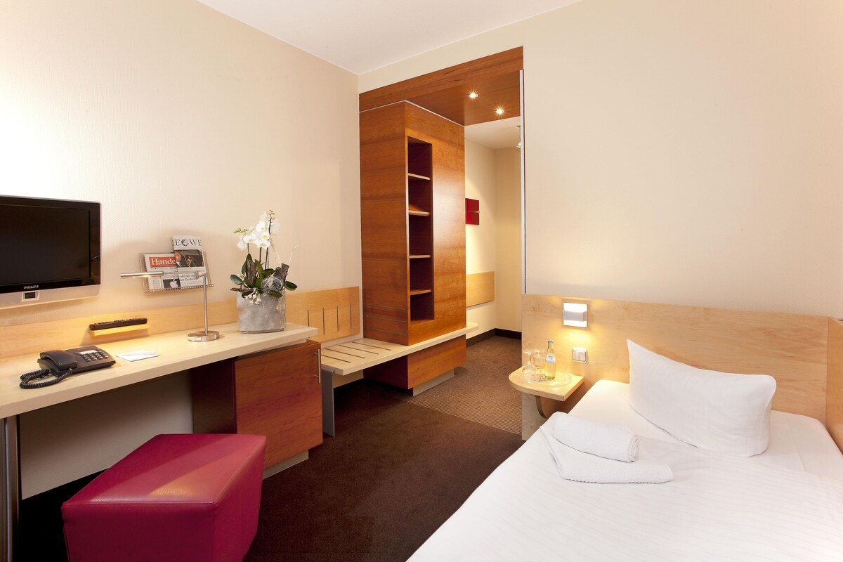 Ein Hotelzimmer mit einem Bett, einem Schreibtisch mit Telefon, einem Spiegel und einem kleinen Tisch mit Flasche und Gläsern.