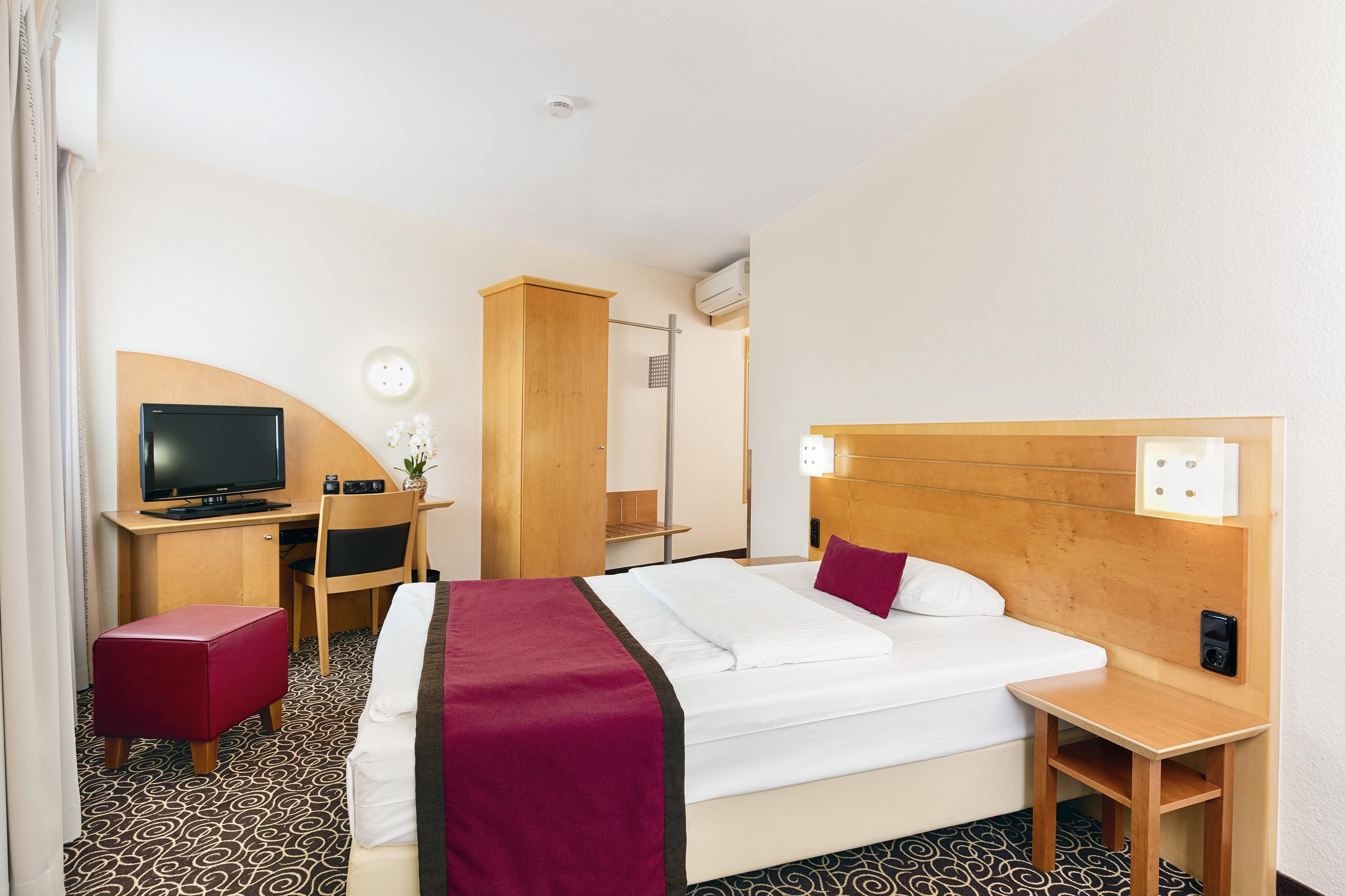 A hotel room with a bed, a desk with a television, a chair, a wardrobe, and a bedside table.