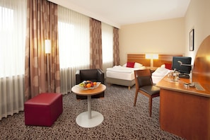 Une chambre d’hôtel avec un lit, un bureau avec un ordinateur, une chaise, une petite table avec des fruits et un pouf rouge.