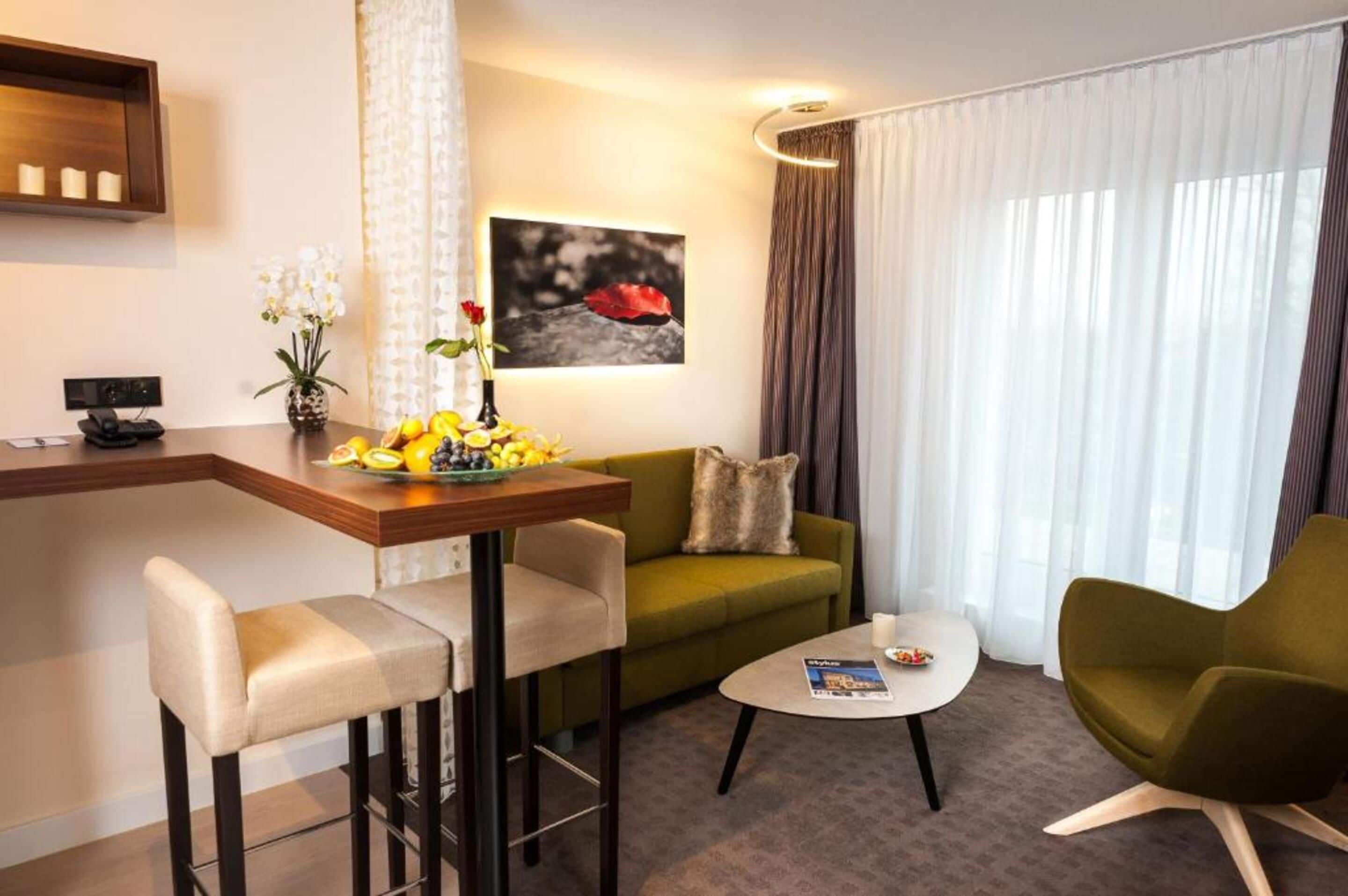 A modern hotel room with a green sofa, a small round table, a bar area with a fruit bowl, and a painting on the wall.
