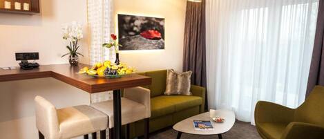 Una habitación de hotel moderna con un sofá verde, una pequeña mesa redonda, una zona de bar con frutero y un cuadro en la pared.
