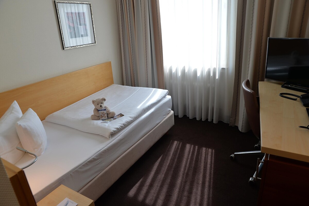 Ein Hotelzimmer mit Bett, Nachttisch mit Fernseher, Teddybär auf dem Bett und Vorhängen.