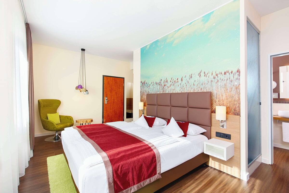 Ein Hotelzimmer mit einem großen Bett, einem grünen Sessel, einem Bild von einem Feld und einem durchgegangenen Tür angedeuteten Badezimmer.