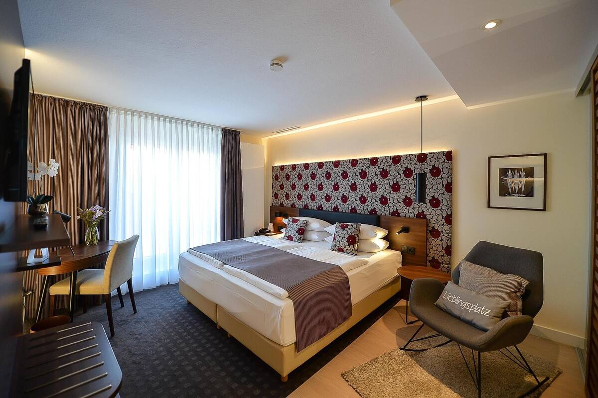 Ein Hotelzimmer mit einem großen Bett, einem Schreibtisch mit Stuhl, einer Couch, einem Fernseher und einem Fenster mit Vorhängen.