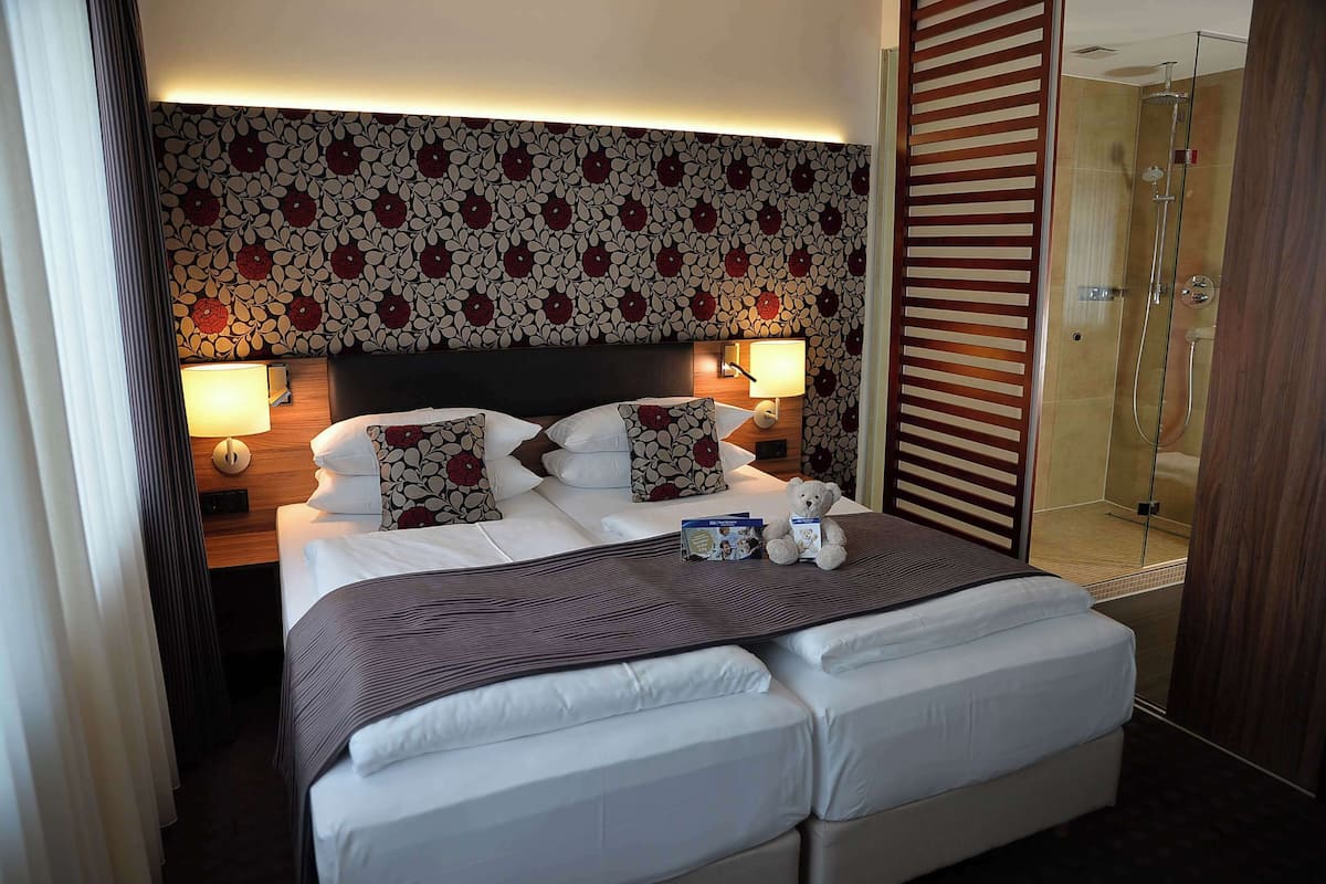 Ein Hotelzimmer mit einem großen Bett, zwei Nachttischen inklusive Lampen, einem blumengemusterten Kopfteil, einer Holztür mit Schieberiegel und einer Duschkabine aus Glas.