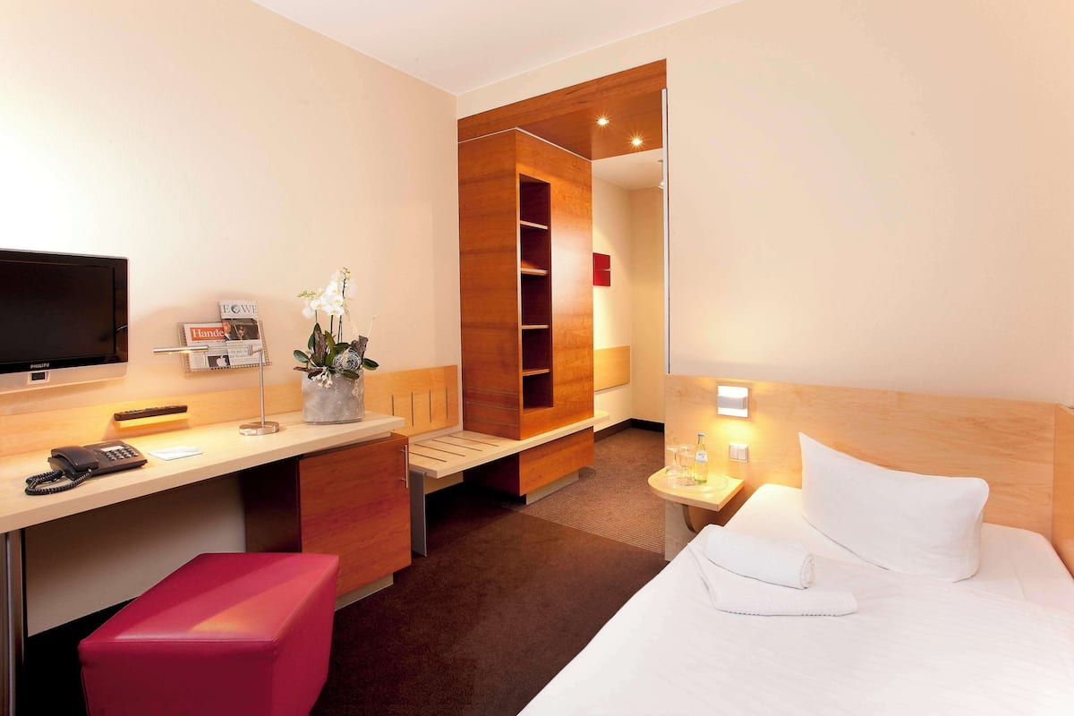 Ein Hotelzimmer mit einem Bett, einem Schreibtisch mit Computer, einem Telefon, einem kleinen Tisch mit Lampe, einer roten Bank und einem Holzschrank.