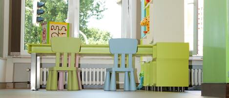 Una habitación luminosa con un ventanal grande, una mesa con sillas y una zona de juegos verde con un juguete de jirafa.