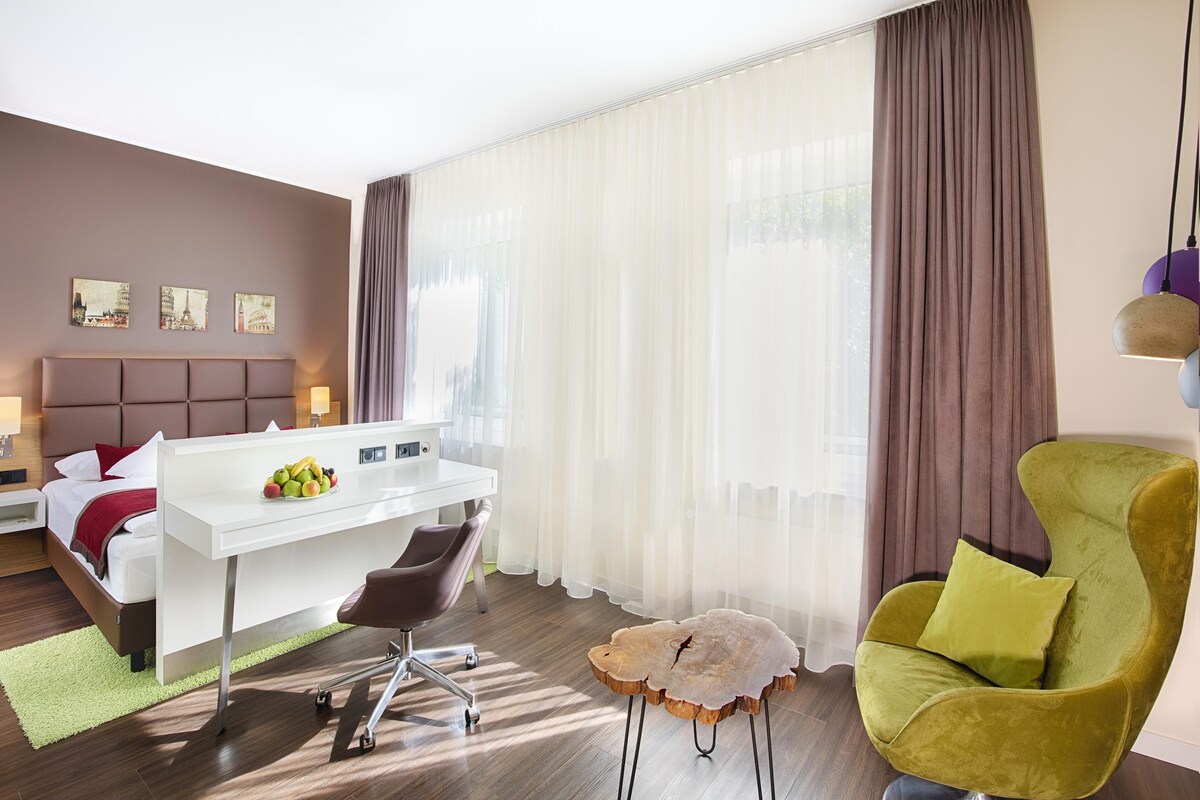 Ein modernes Hotelzimmer mit einem Bett, einem Schreibtisch, einem Sessel und einem großen Fenster mit Vorhängen.