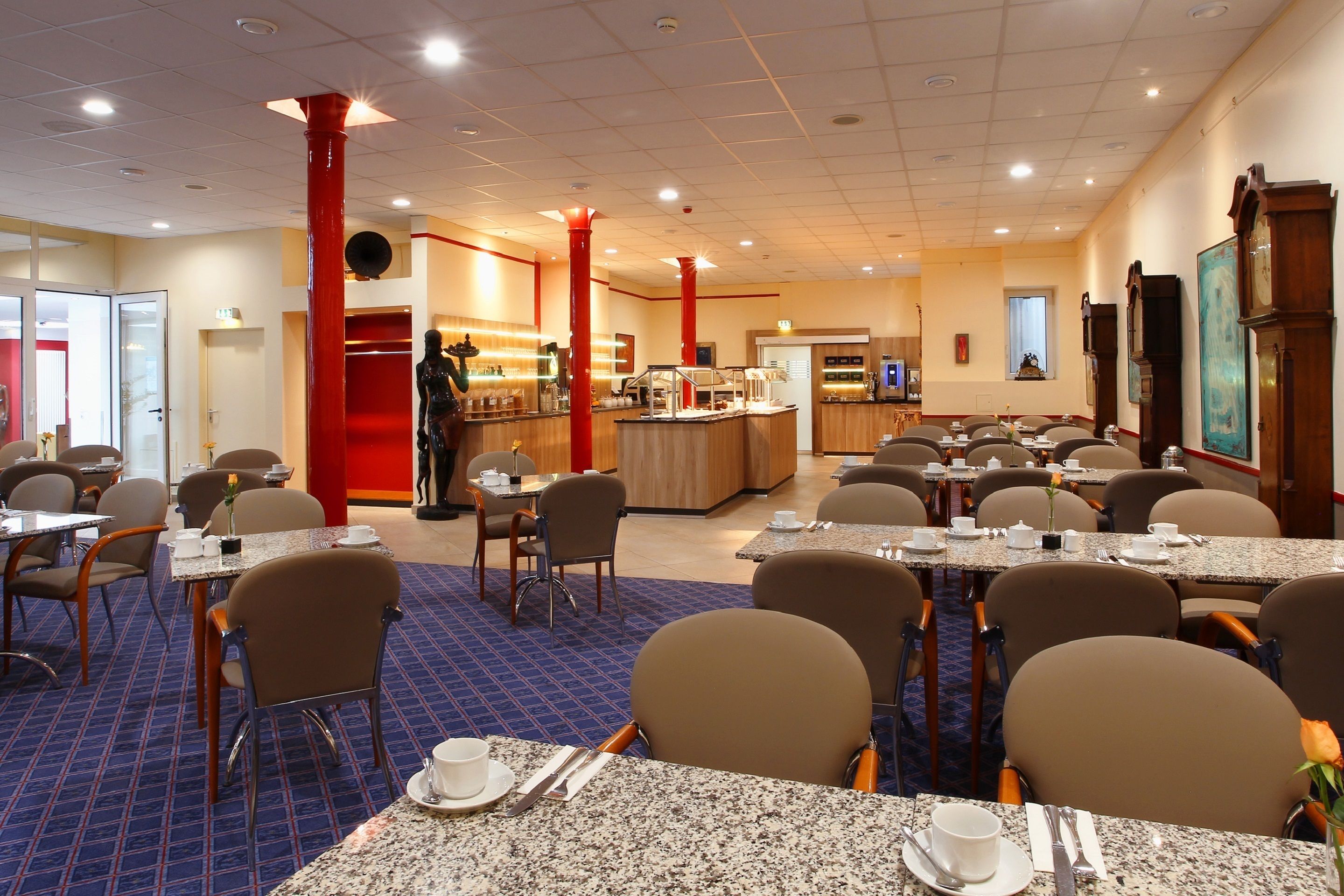 Comedor de hotel con varias mesas y sillas, una barra central con una persona atendiendo, y una zona de cocina al fondo.