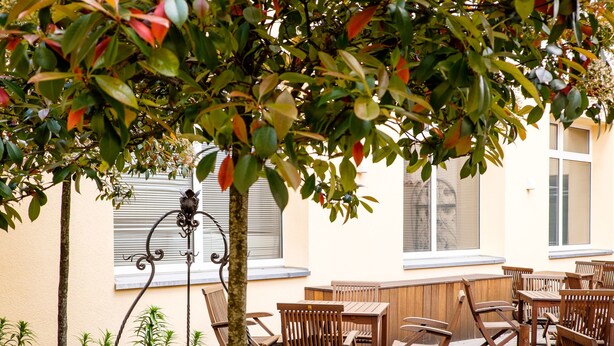 An outdoor seating area with wooden tables and chairs, surrounded by lush greenery and a decorative metal stand.