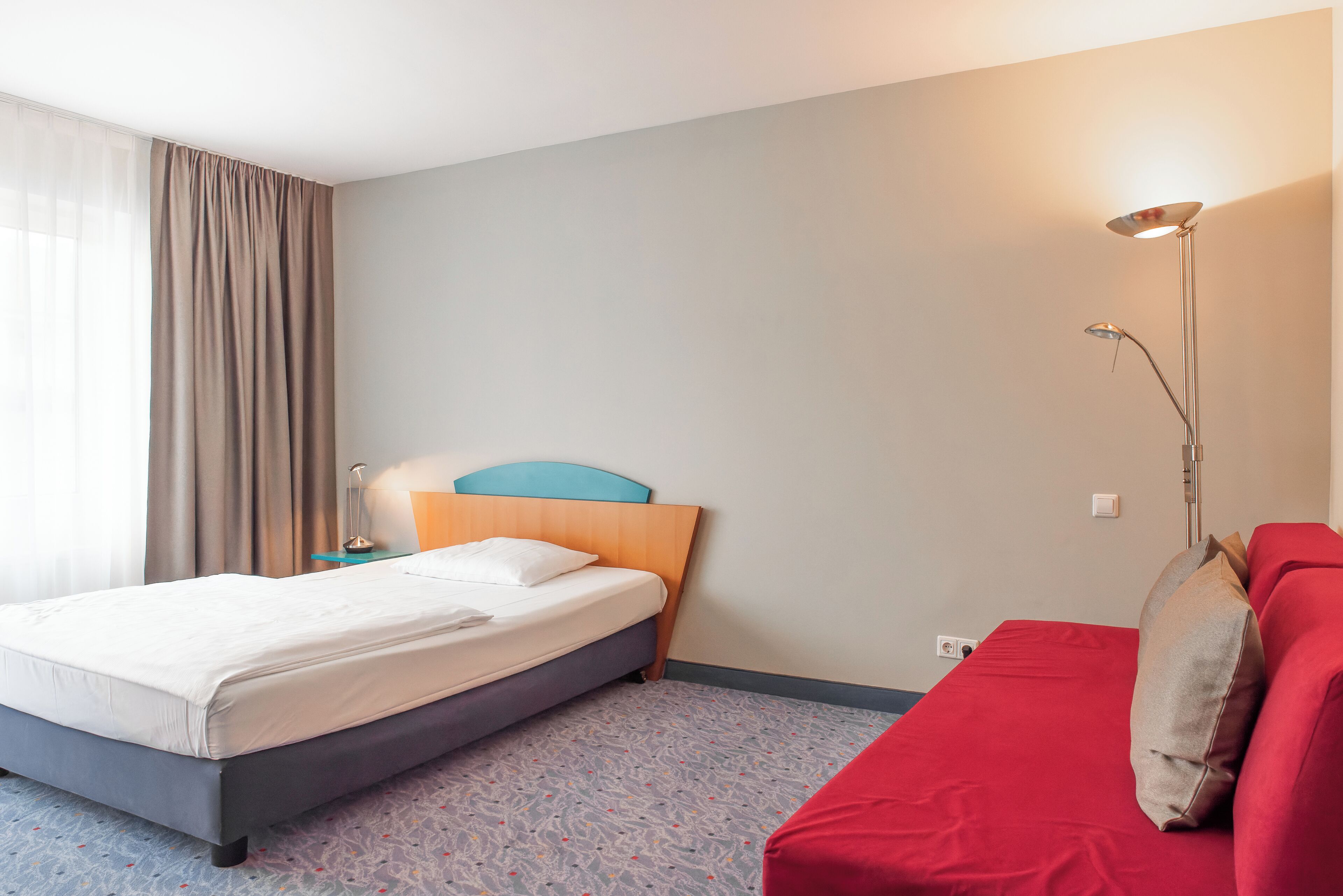 Una habitación de hotel con cama, un sofá rojo, una lámpara y una ventana con cortinas.