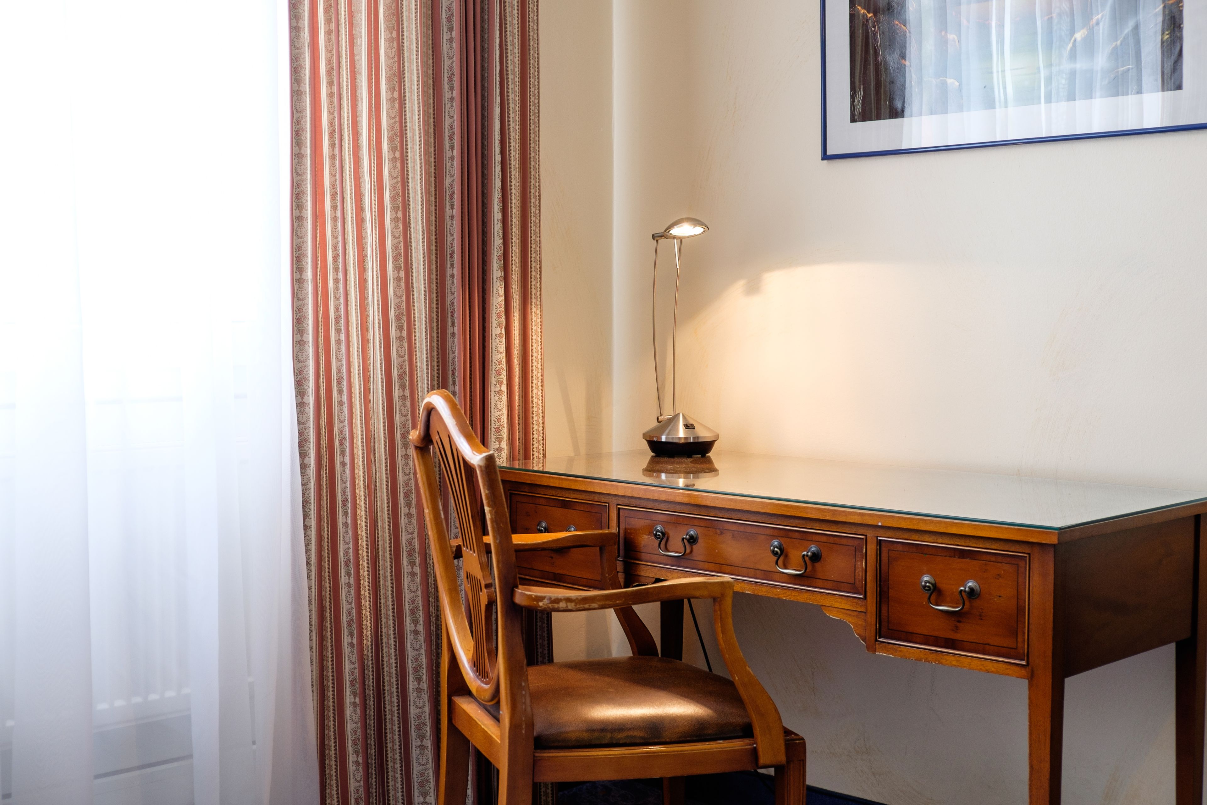 A wooden desk with a chair, a lamp, and a framed picture on the wall.