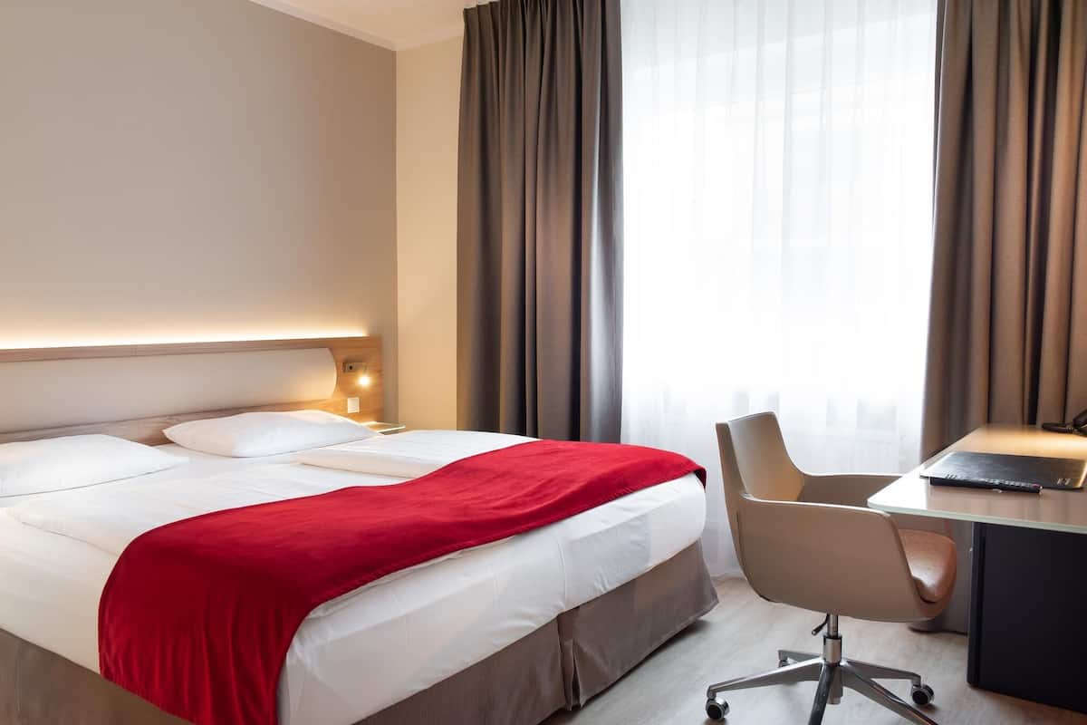 Ein Hotelzimmer mit Bett, Schreibtisch, Stuhl und einem fenster mit Vorhängen.