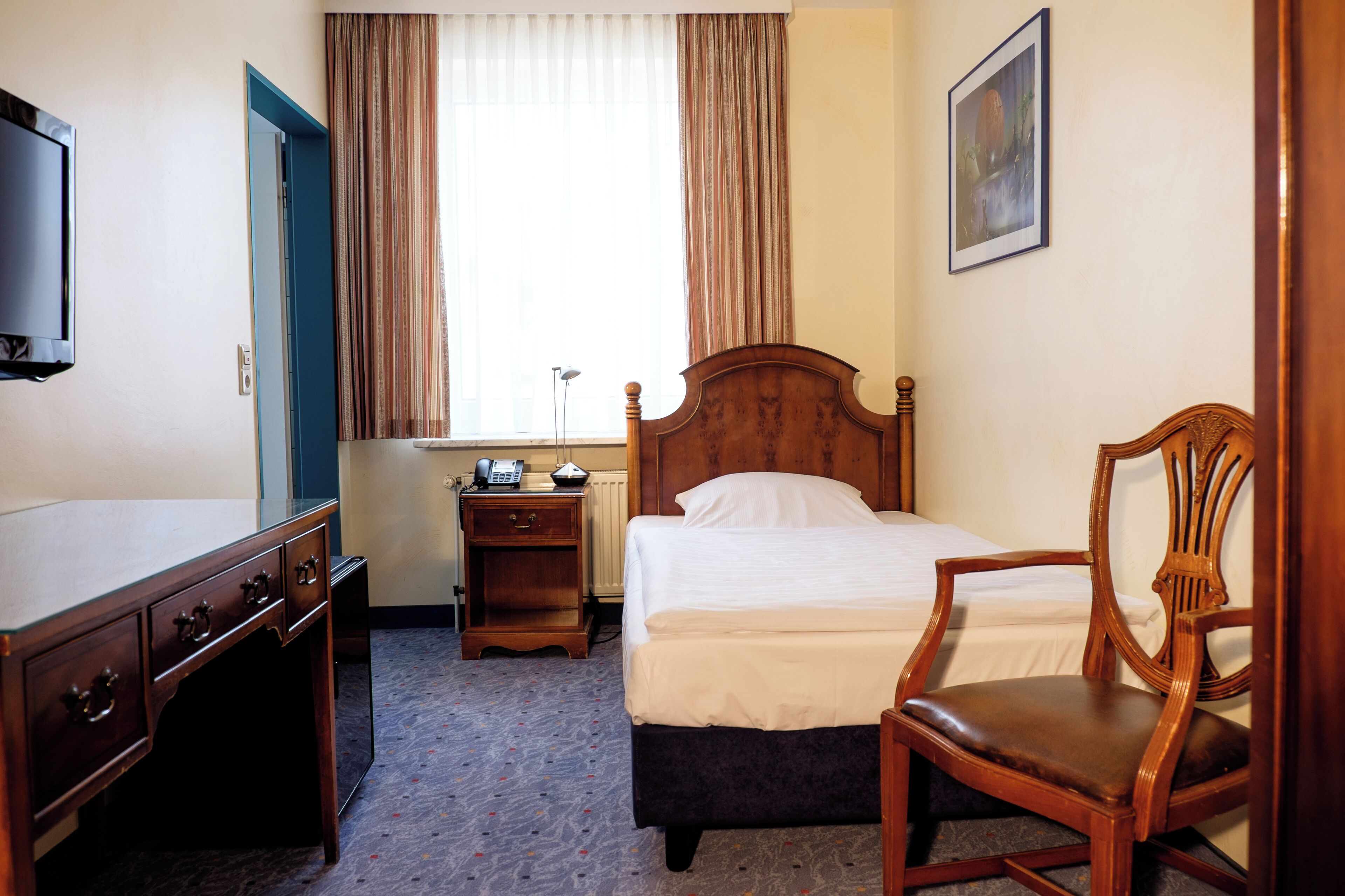 A hotel room with a bed, a wooden chair, a desk with drawers, a bedside table with a lamp, and a painting on the wall.