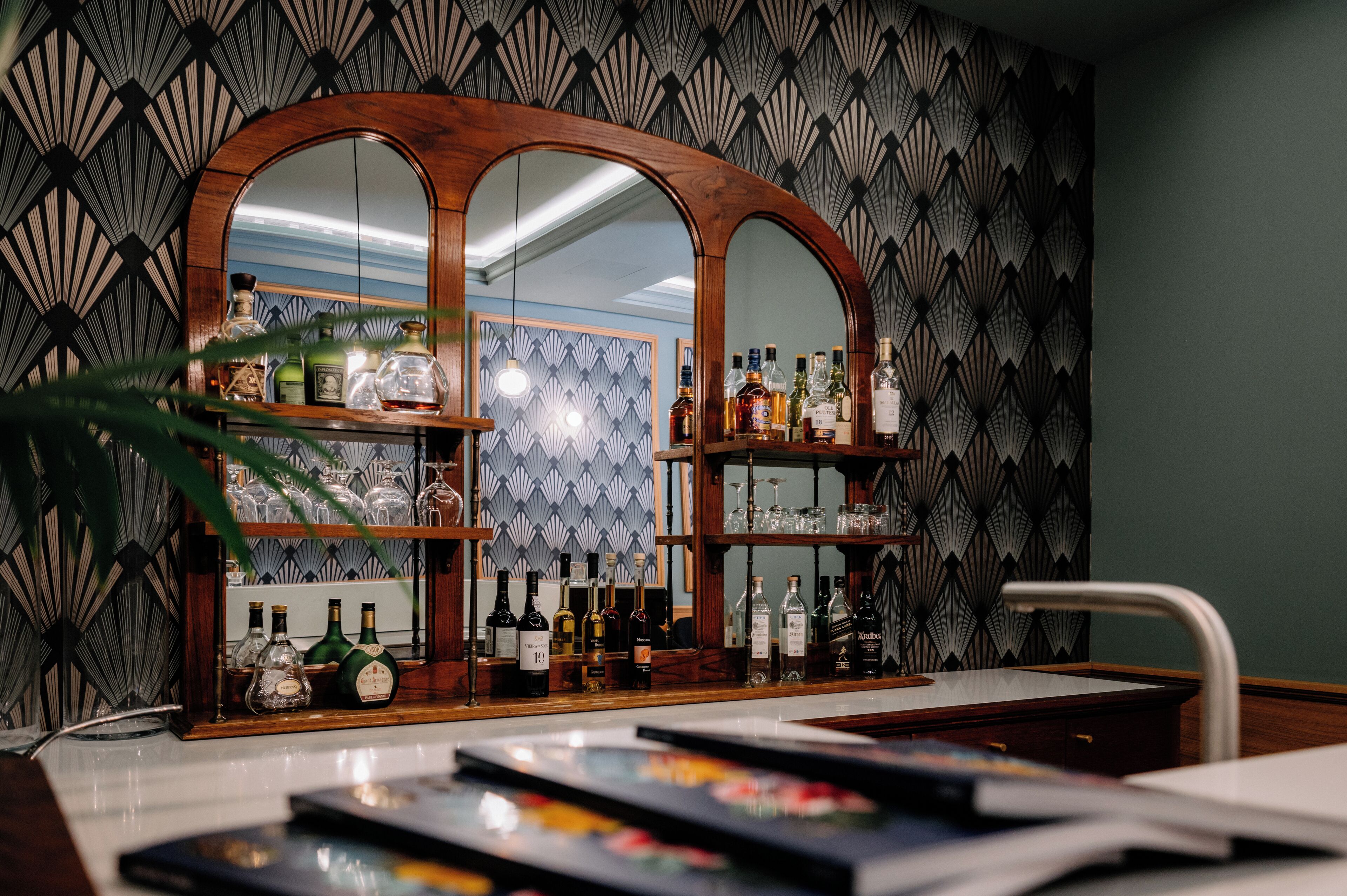 Un coin bar agrémenté d’un miroir en bois, d’étagères remplies de bouteilles et offrant une vue sur une pièce dont les murs sont recouverts d’un papier peint à motifs.