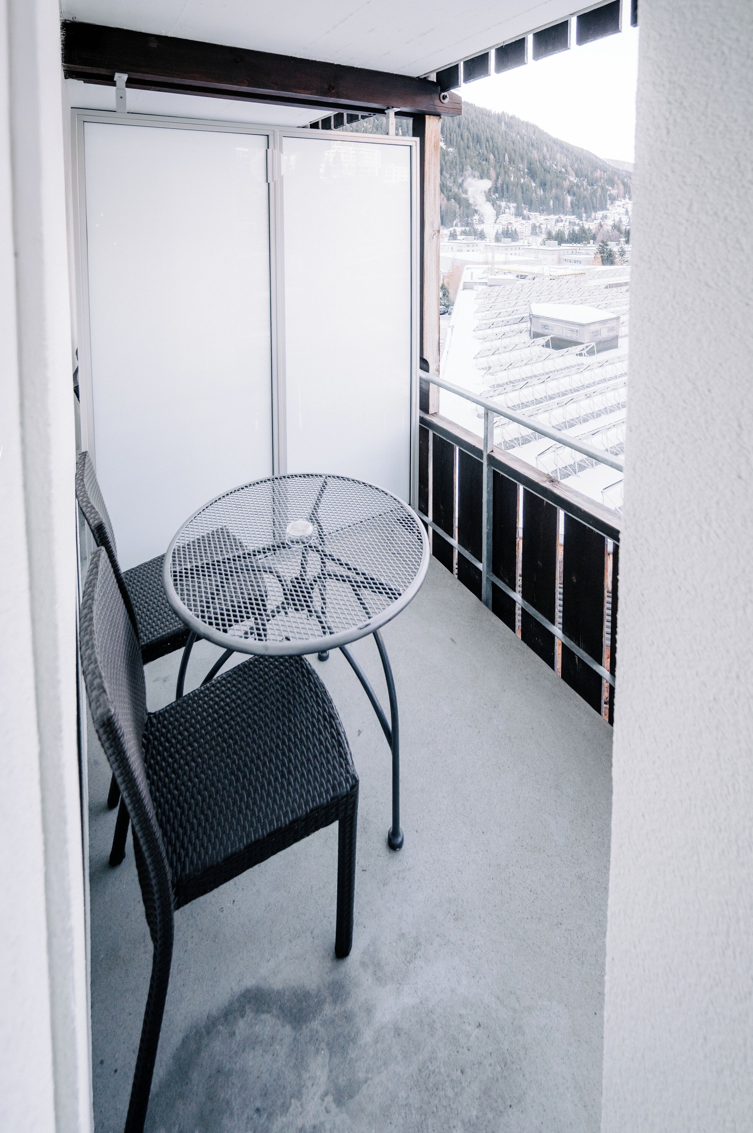 A balcony with a metal table and two wicker chairs, overlooking a snowy landscape with buildings and a mountain.