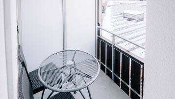 A balcony with a metal table and two wicker chairs, overlooking a snowy landscape with buildings and a mountain.