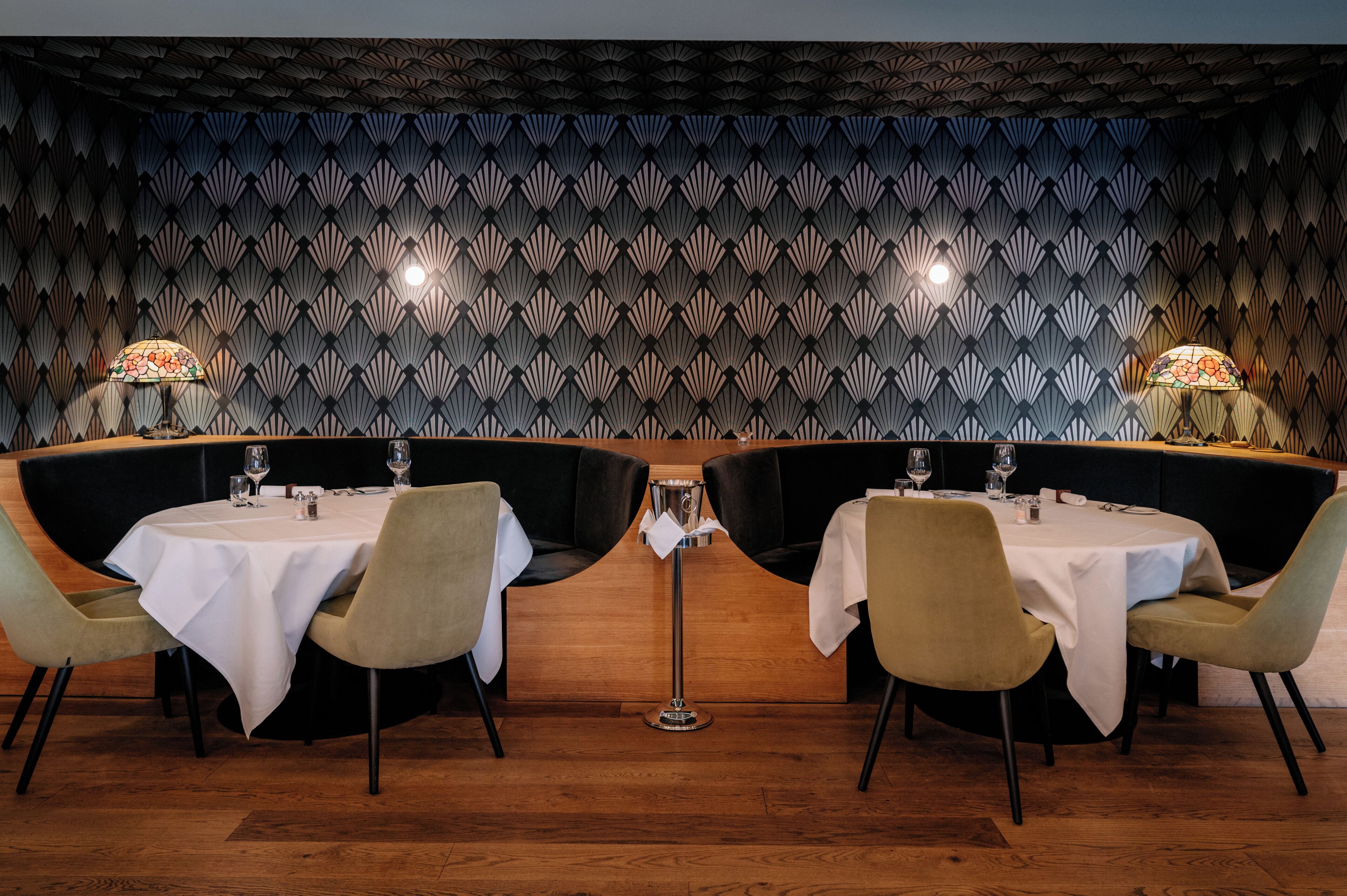Ein Restaurant mit zwei runden Tischen, jeder mit einer weißen Tischdecke und vier Stühlen. Die Wände weisen ein geometrisches Muster auf, und auf jedem Tisch stehen zwei Tischlampen.
