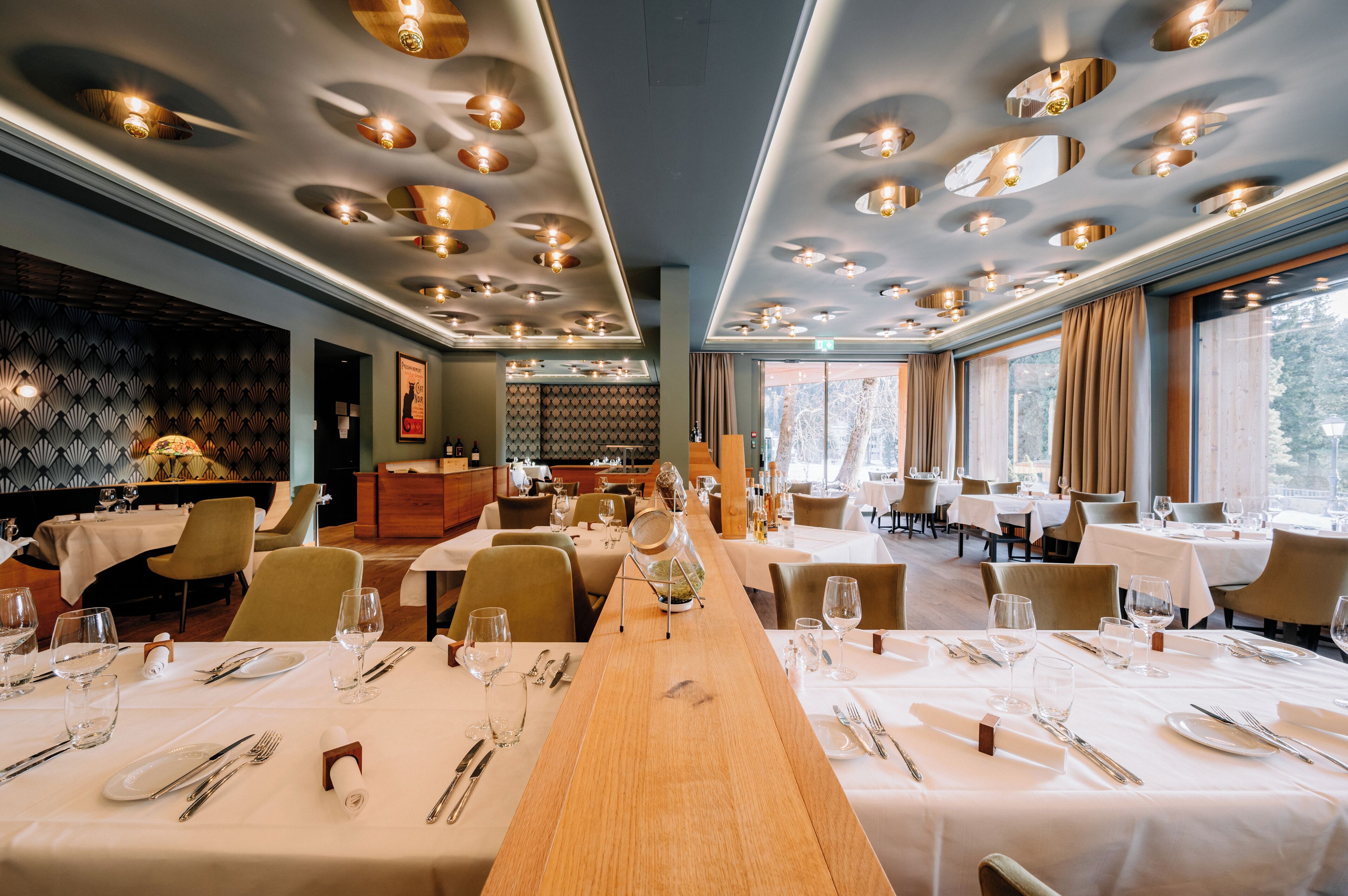 Un restaurante con varias mesas dispuestas para comensales, un área de cocina visible al fondo y amplias ventanas que dejan entrar luz natural.
