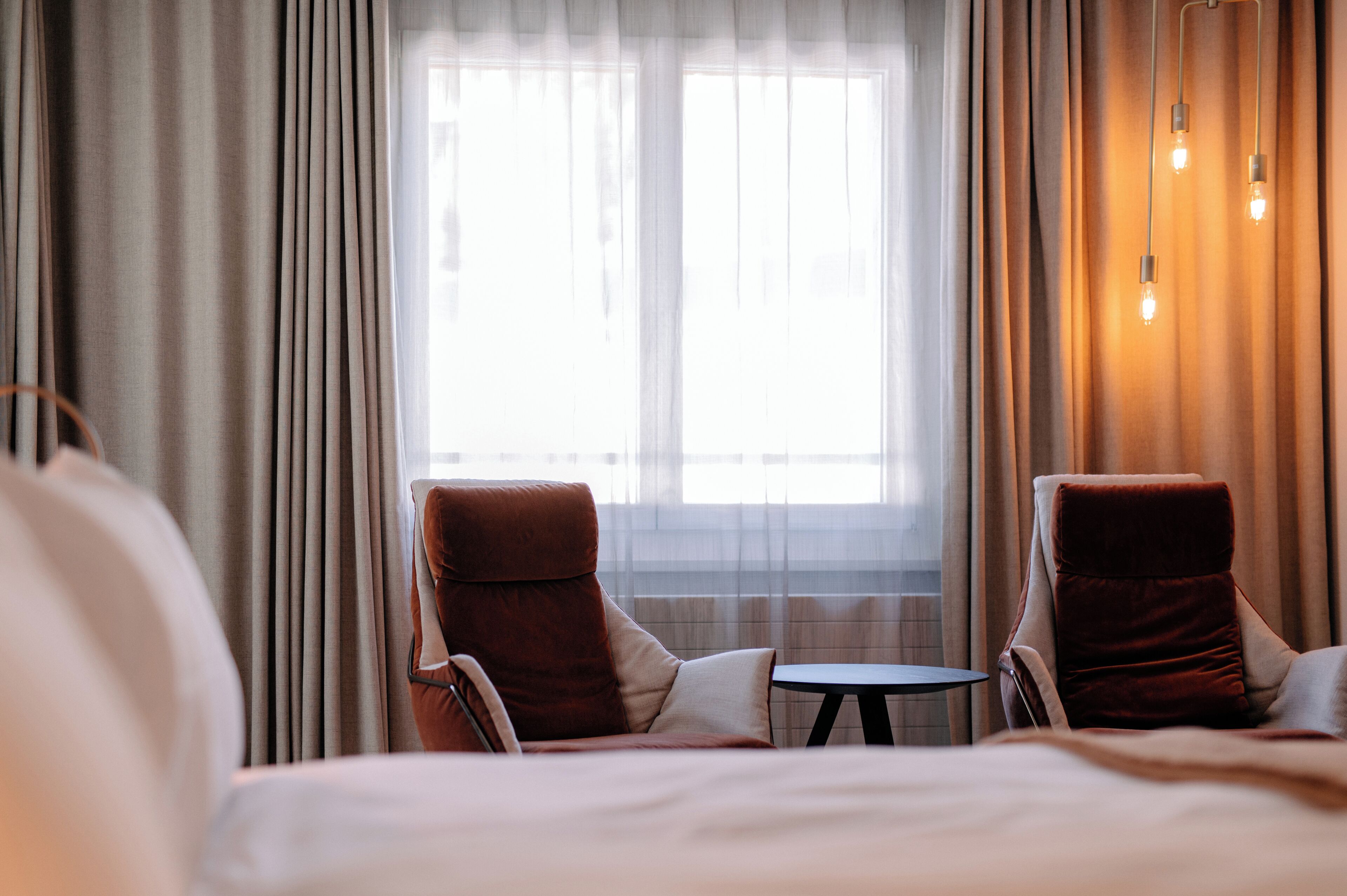A hotel room with a bed, two armchairs, a small table, and a window with curtains.