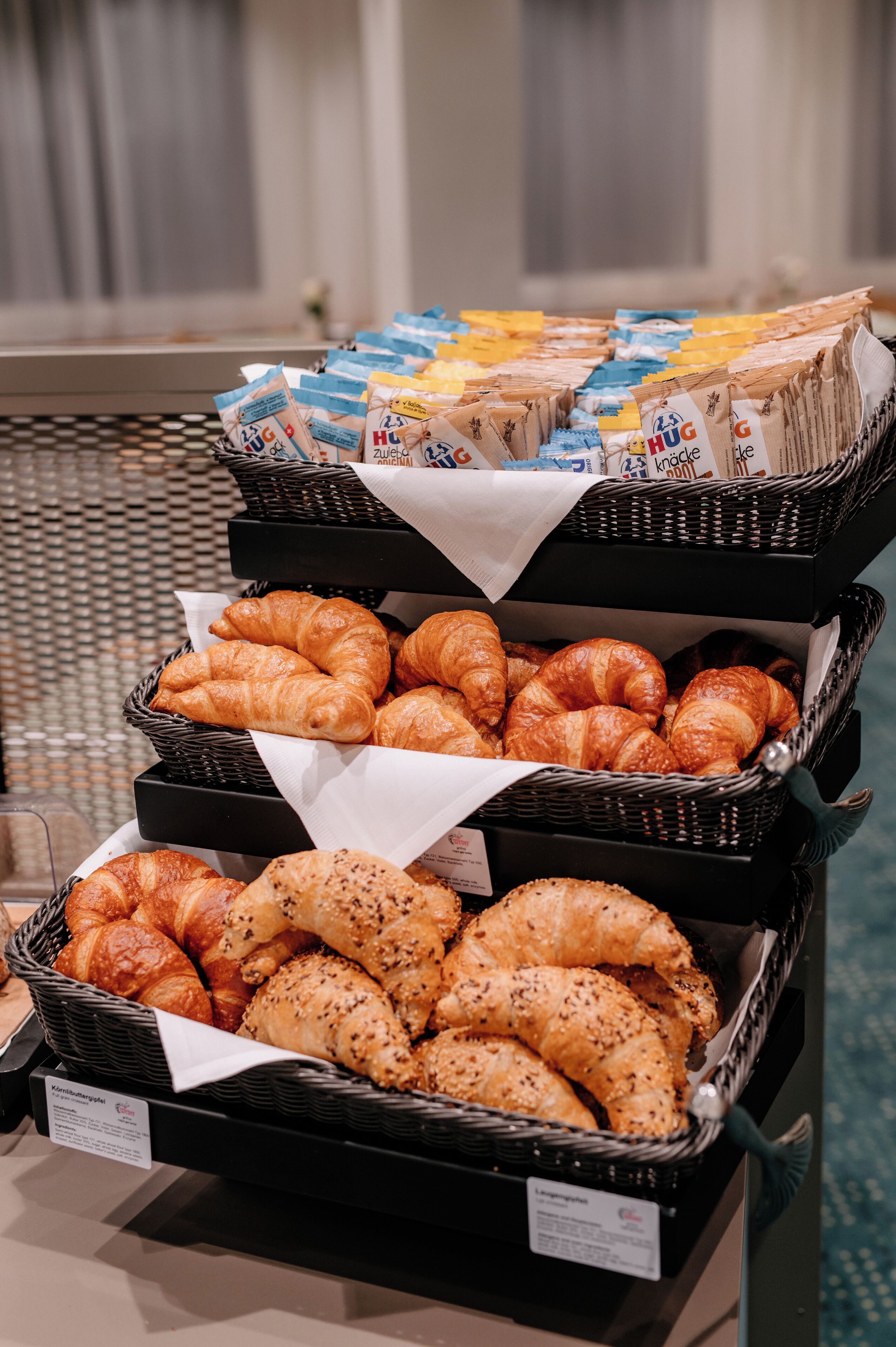 Exposição de diversos pastéis em cestos de vime, incluindo croissants e bagels, com etiquetas de preço visíveis.