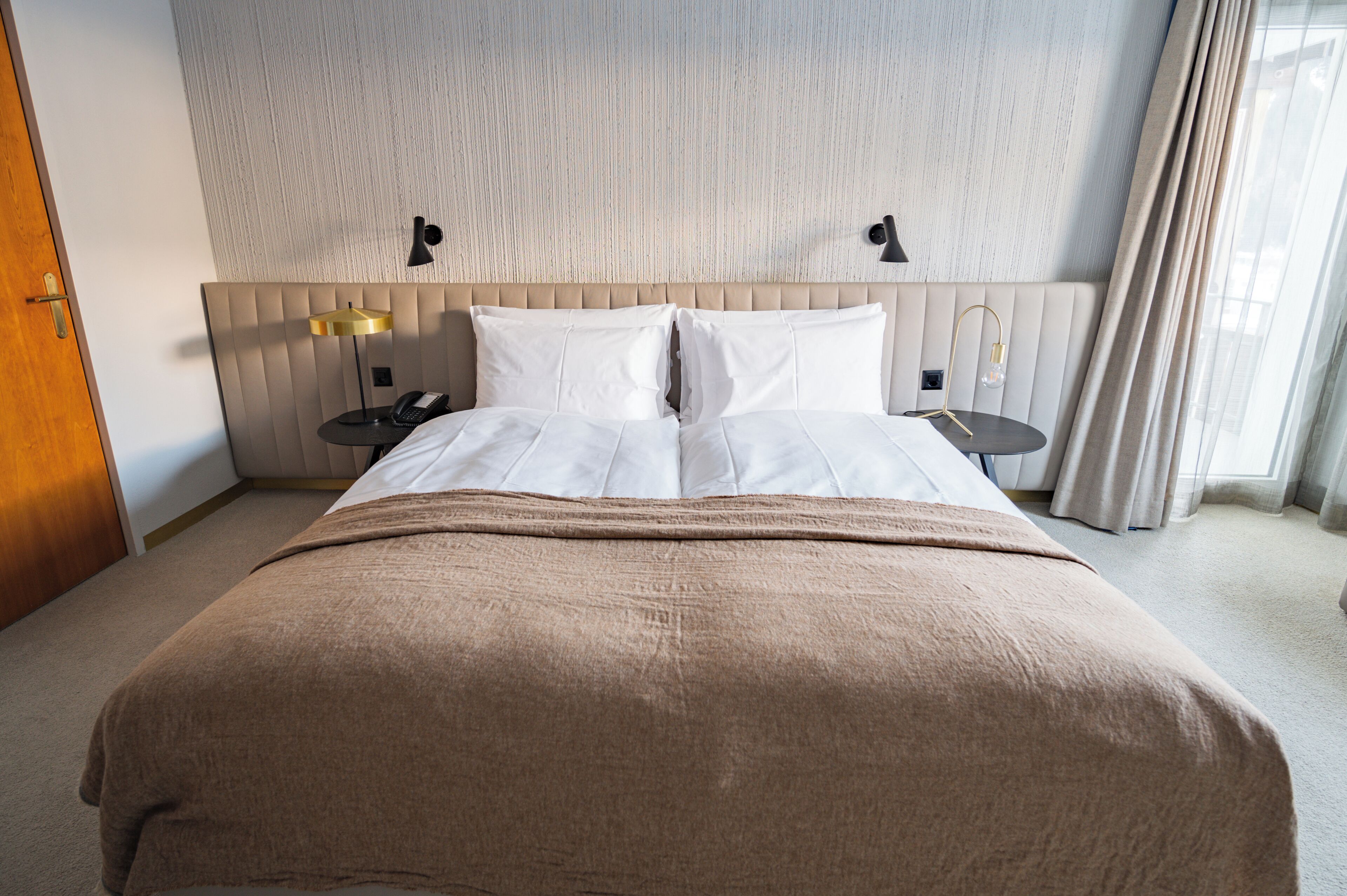 A hotel room with a large bed, a headboard with vertical lines, two bedside tables with lamps, and a door on the left.