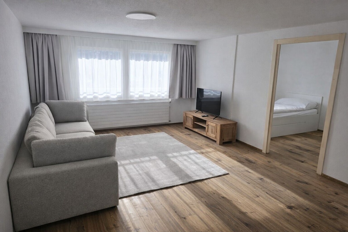 A modern living room with a grey sofa, a wooden TV stand, and a bed in the adjacent room.
