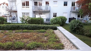 Ein Wohngebäude mit weißer Fassade, mehreren Balkonen und einem gepflegten Garten mit Sträuchern und einem Kiesweg.