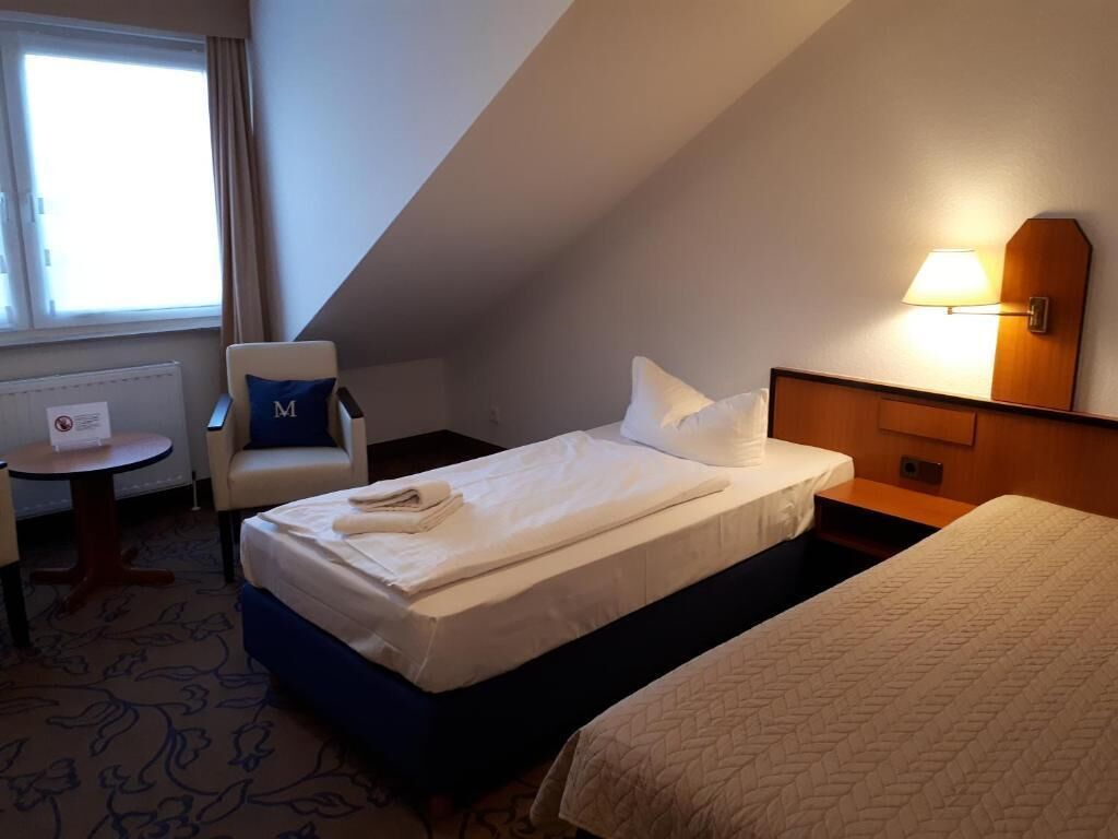 A hotel room with a single bed, a chair with a pillow, a small table, and a lamp.
