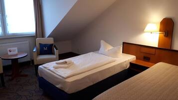 A hotel room with a single bed, a chair with a pillow, a small table, and a lamp.