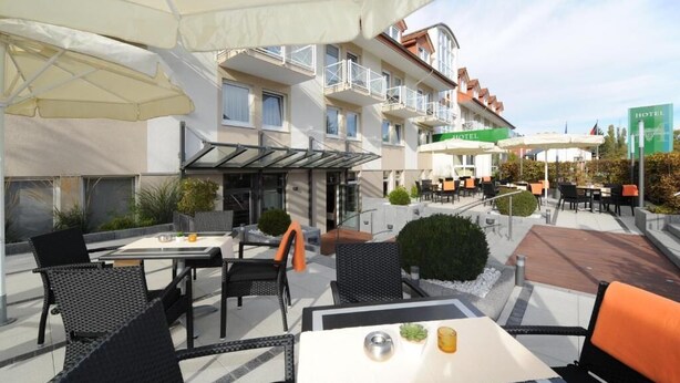 Ein Außenbereich mit Terrasse, Tischen, Stühlen und Sonnenschirmen, sowie ein Gebäude mit Balkonen.
