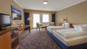 Ein Hotelzimmer mit zwei Betten, einem Schreibtisch mit Fernseher, einem Sessel, einem kleinen Tisch und einem Fenster mit Jalousien.