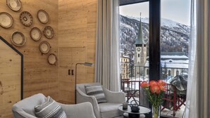 A cozy room with two grey armchairs, a small table, and a vase with flowers. A large window offers a view of a snowy mountain and a church. - Unique Hotel Post (Zermatt)