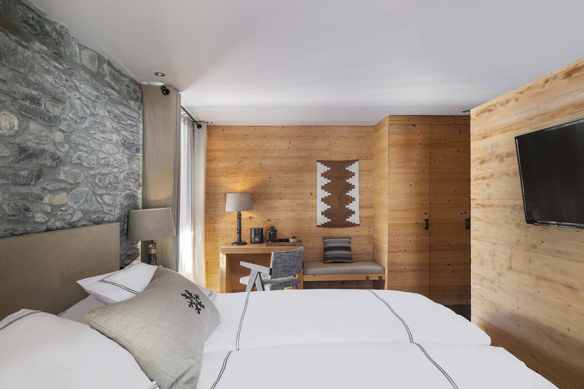 A hotel room with a stone wall, wooden paneling, a bed with white linens, a desk with a lamp, and a TV mounted on the wall.