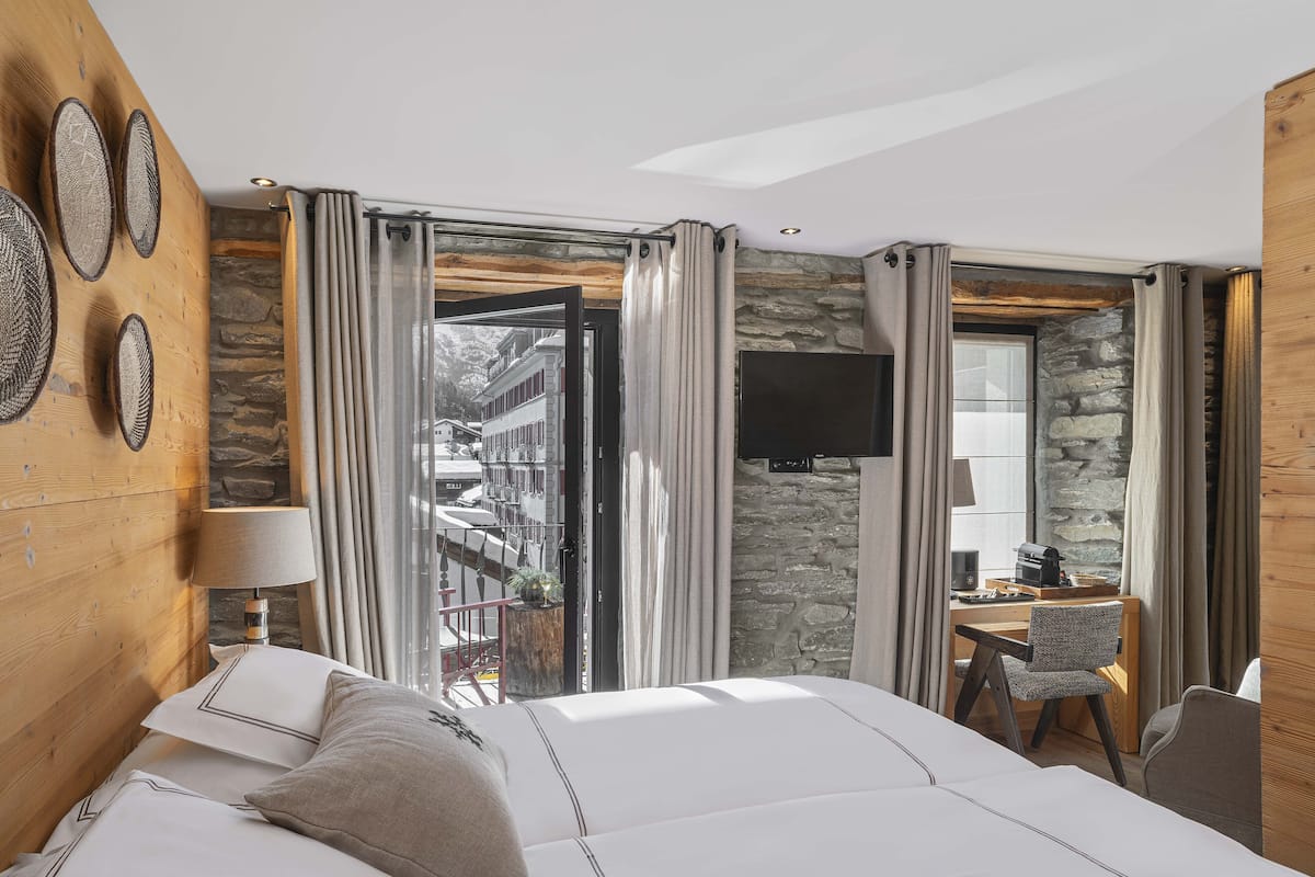 A hotel room with a bed, a desk with a chair, a TV, and a stone wall.