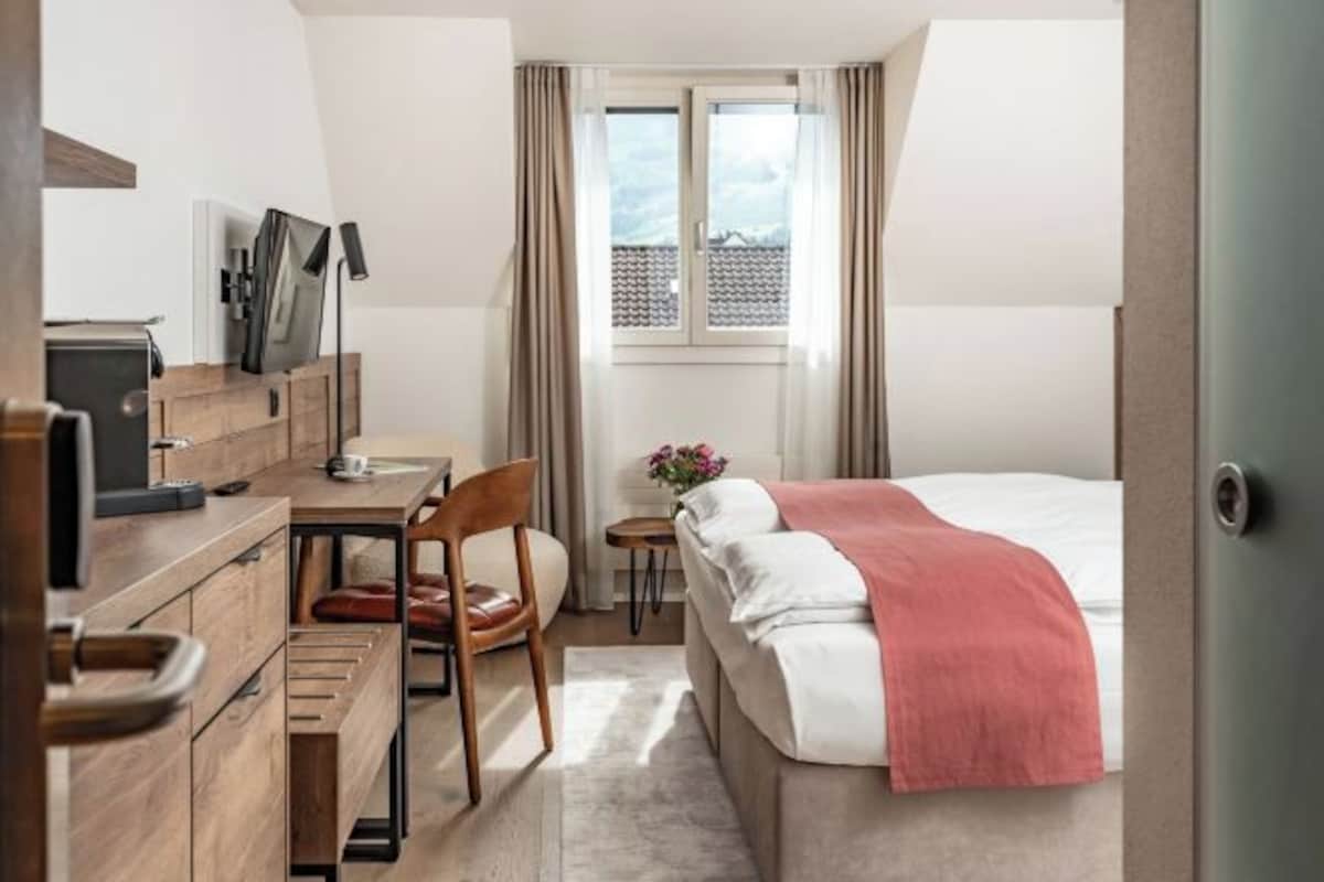 A hotel room with a bed, a desk with a chair, a small table with a vase of flowers, and a window with curtains.