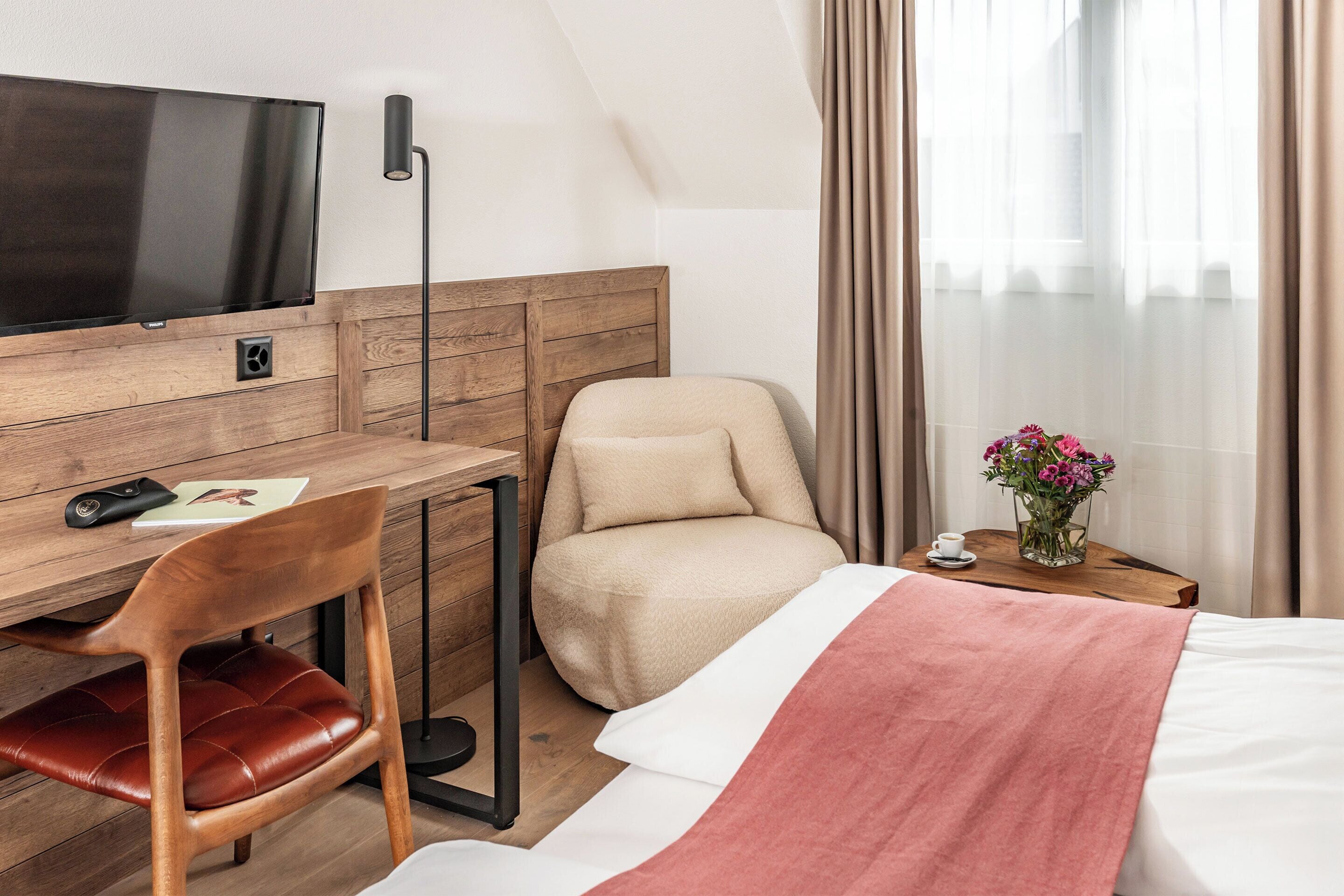Habitación de hotel con escritorio de madera, televisor, una butaca cómoda, cama con manta rosa y una mesita con un jarrón de flores.