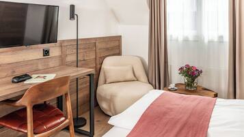 Habitación de hotel con escritorio de madera, televisor, una butaca cómoda, cama con manta rosa y una mesita con un jarrón de flores.