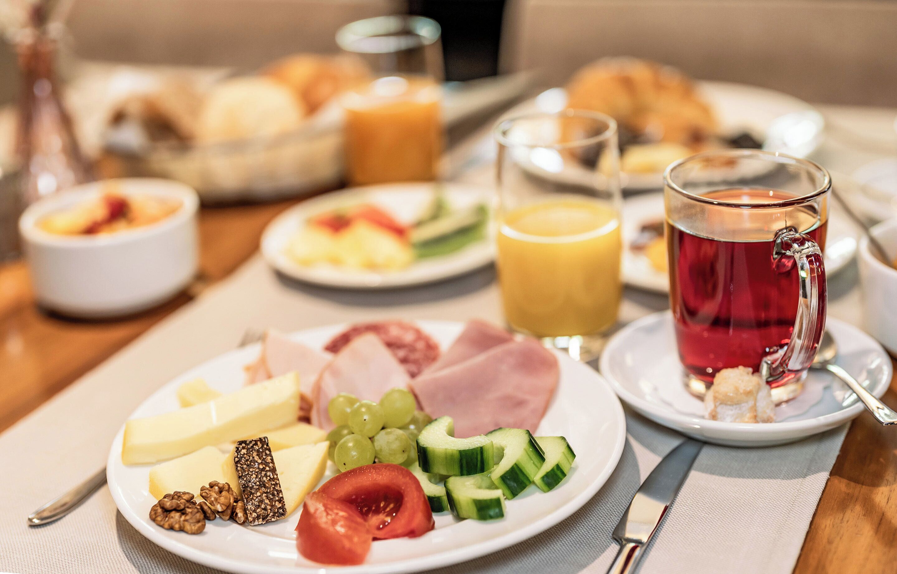 Una mesa de desayuno con platos de queso, jamón, uvas y verduras, un vaso de jugo y una taza de té.
